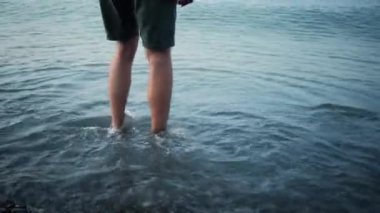 Şortlu beyaz adam, çakıl taşı kumsalının sığ sularında ayak bileğine kadar suyun içinde duruyor. 4K yavaş çekim videosu suyun içindeki ayakları yakından gösteriyor. Küçük deniz dalgaları çıplak bacakların üzerinde yuvarlanıyor..