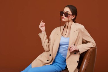 Bej ceketli ve kot pantolonlu zarif bir kadının yüksek moda portresi. Güneş gözlüğü, kahverengi arka plan, stüdyo çekimi