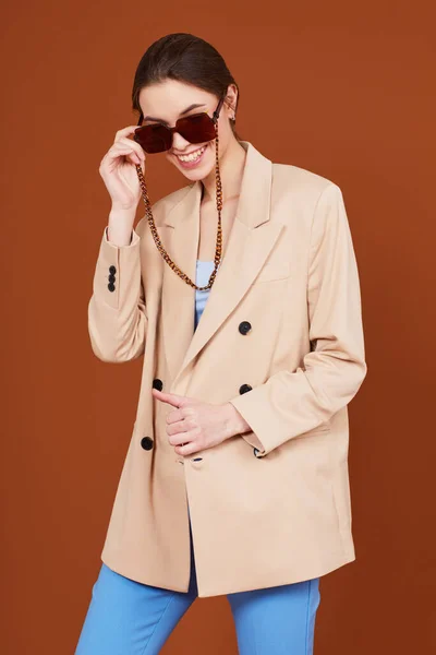 ベージュのジャケットとジーンズで若いエレガントな女性の高いファッションの肖像画 サングラス 茶色の背景 スタジオショット — ストック写真