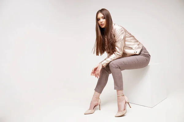 Fashion model in golden leather jacket posing in studio. Brunette hair, brown pants, shoes