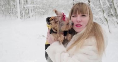 Beyaz kürklü güzel bir kadın Yorkshire Teriyeri 'nin küçük köpeğiyle karlı ormanda poz verirken kameraya bakıyor. Yorkie sahibini yalıyor.  