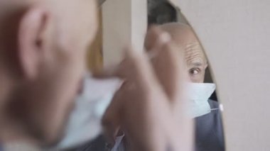 Yaşlı adam aynadaki yansımaya bakıyor ve koruyucu yüz maskesi takıyor.