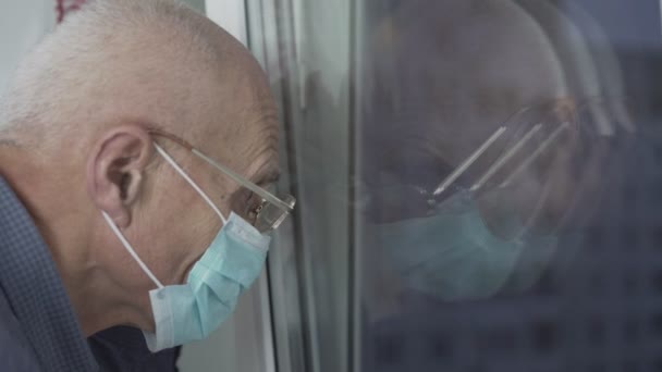 Senior man draagt beschermende gezichtsmasker kijken door het raam glas in de avond — Stockvideo