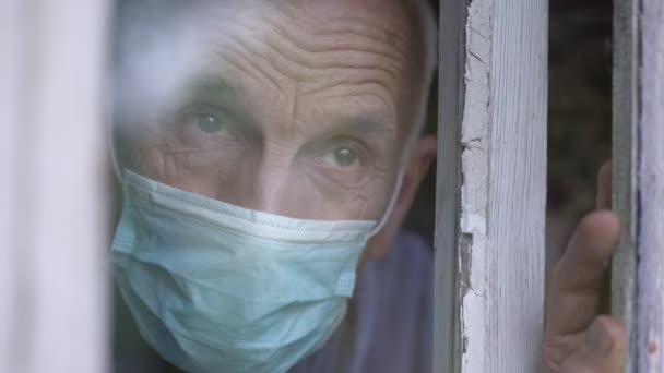 Aged man in protective facemask looking with sorrow through window glass — Stock Video