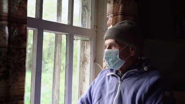 Viejo agricultor con gorra de calidez y mascarilla médica mirando a través de la ventana — Vídeos de Stock