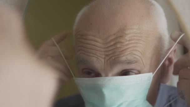 Ηλικιωμένος συνταξιούχος με ρυτιδιασμένο πρόσωπο που τίθεται σε ιατρική facemask κοιτάζοντας τον καθρέφτη — Αρχείο Βίντεο