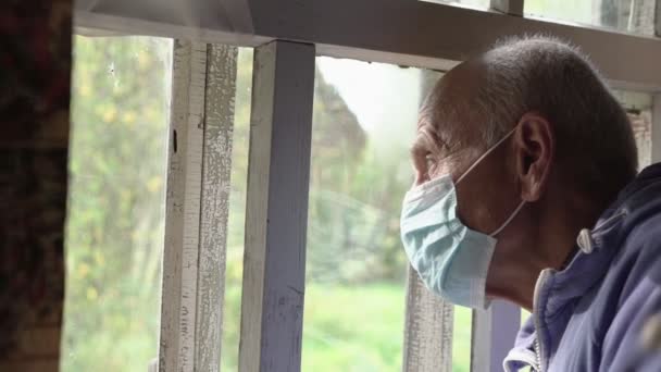 Пожилой лысый мужчина носит медицинскую синюю маску, смотрит в окно деревенского дома — стоковое видео