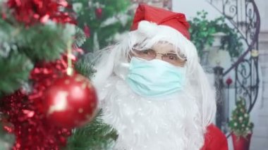 Yüz maskeli ve kırmızı şapkalı komik Noel Baba Noel ağacının yanında eğleniyor.