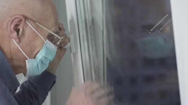 Hombre mayor en gafas y mascarilla médica saludando con la mano a través de la ventana — Vídeos de Stock