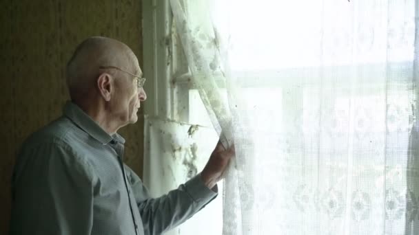 Leeftijd man draagt brillen houdt wit gordijn met de hand en kijkt door het raam. — Stockvideo