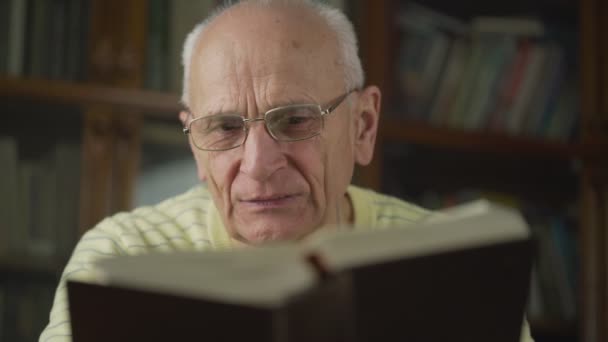 Elderly man wearing eyeglasses reads book carefully close view indoor — Stock Video