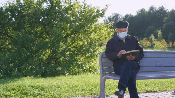 Homem sênior em máscara protetora sentado no banco e lendo livro vintage — Vídeo de Stock