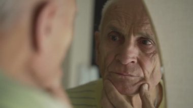 Üzgün yaşlı adam banyo aynasına bakıyor ve parmaklarıyla tenine dokunuyor..