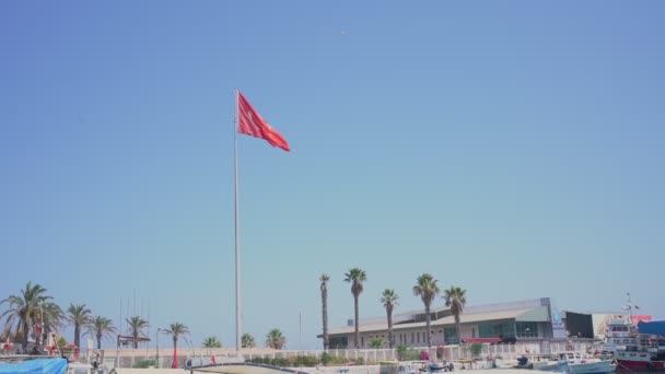 Bandera roja turca ondeando en cielo azul claro bajo muelle urbano con barcos de pesca. — Vídeo de stock