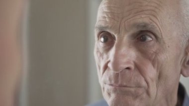 Yaşlı adam aynaya bakıyor ve yüzüne mavi tıbbi maske takıyor..