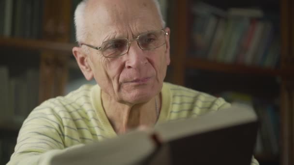 眼鏡をかけた高齢の白髪の男は注意深く大きな本を読んでいます. — ストック動画