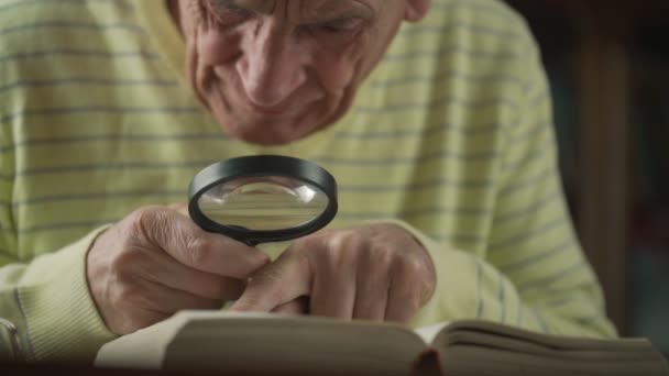 Gammal rynkig man med förstoringsglas i händerna läser bok — Stockvideo