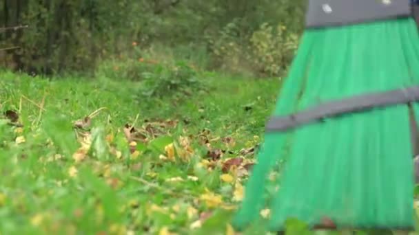 Gardener in rubber boots sweeps with green broom fallen autumn foliage — Stock Video