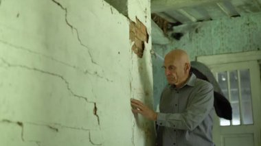 Kel yaşlı bir adam kırsal kesimdeki eski bir evde beyaz çatlamış duvarın yanında duruyor..