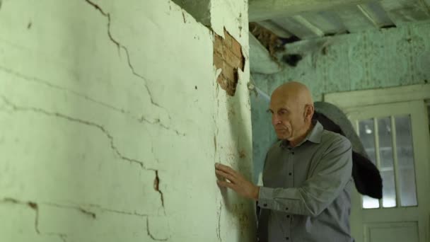 Kale oudere man staat in oude landelijke huis in de buurt van witte gebarsten muur. — Stockvideo