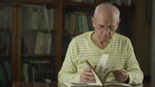 Gammal professor i glasögon sittande vid bord och blad genom gammal bok. — Stockvideo