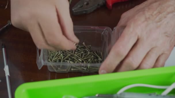 Homem com as mãos enrugadas tirando parafusos de metal do recipiente de plástico na mesa. — Vídeo de Stock