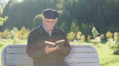 Mature male citizen wearing cap and glasses sitting on bench and reads book.