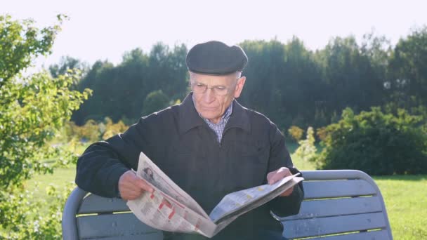 Åldrad man i glasögon läser tidningen sitter på bänk i parken — Stockvideo