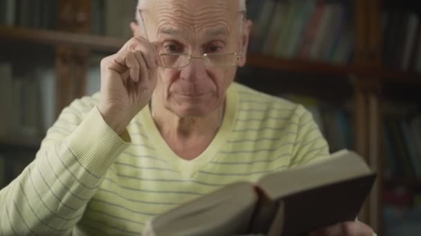Втомлений старий у окулярах шукає відповіді у релігії з біблійною книгою в руках.. — стокове відео