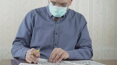Tıbbi maskeli yaşlı bir adam gazete bulmacasında kelimeler yazıyor..