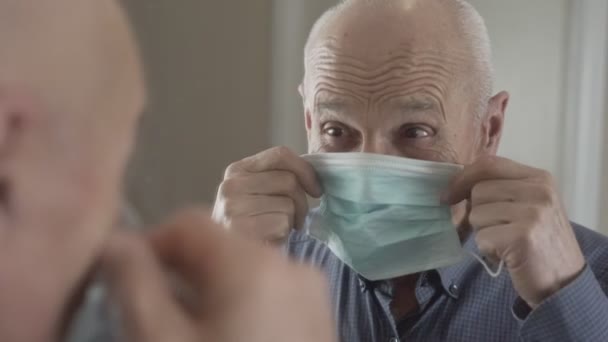 Az idősebb férfi orvosi maszkot vesz fel az arcára, és tükörbe néz.. — Stock videók