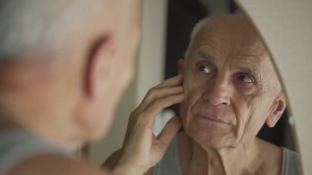 Pria berambut abu-abu menyentuh wajah keriput dengan tangan melihat di cermin — Stok Video