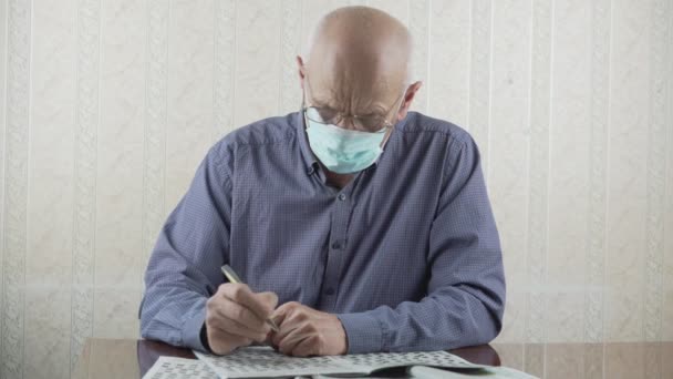 Oudere kale persoon in beschermend gezichtsmasker slovend kruiswoordraadsel met pen — Stockvideo
