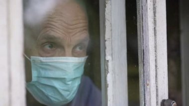 Pencereden bakan, tıbbi maske takan yaşlı bir erkek..