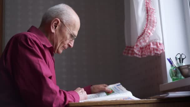 Oude grijze grootvader zit aan tafel en leest krant. — Stockvideo