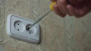 Elinde tornavida olan bir adam, duvardaki beyaz plastik soketten vidaya dönüşüyor..