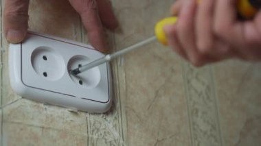 Yaşlı adam oda duvarındaki beyaz prizi tornavidayla onarıyor..