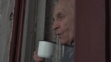 Üzgün yaşlı bir adam pencere camının arkasında durur ve bardaktan çay içer..