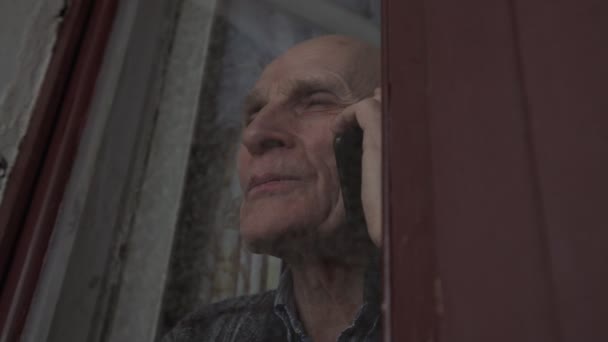 Пожилой человек стоит за стеклом и звонит по смартфону. — стоковое видео
