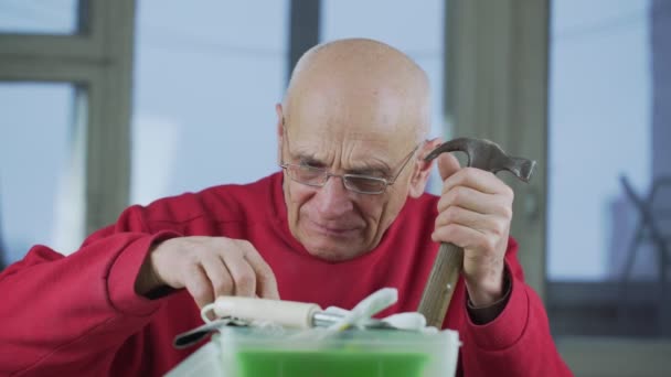 안경을 쓴 노인 이 식탁에 앉아 손으로 망치와 렌치를 쥐고 있다 — 비디오