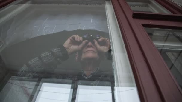 Loco anciano hombre sostiene los prismáticos en las manos y mirando a través de la ventana — Vídeo de stock