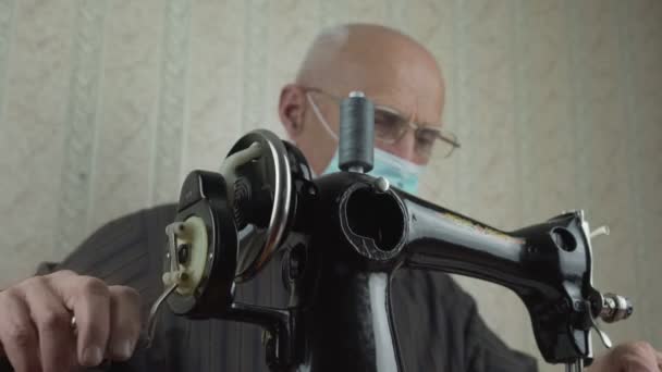 Senior man met bril en gezichtsmasker naaien met vintage machine thuis. — Stockvideo