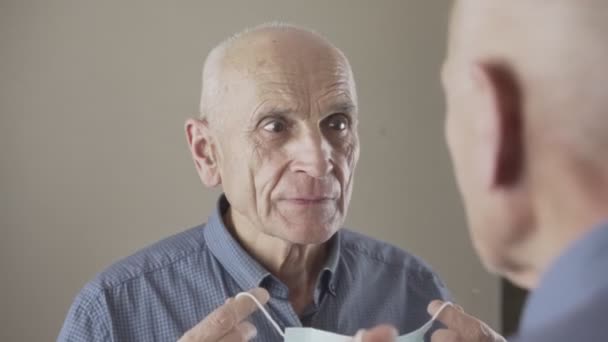Yaşlı beyaz adam aynaya bakıyor ve tıbbi maske takıyor. — Stok video