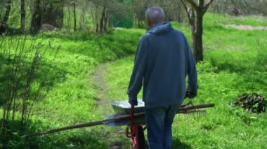 Kapüşonlu yetişkin çiftçi bahçeye tarımsal aletlerle giriyor.