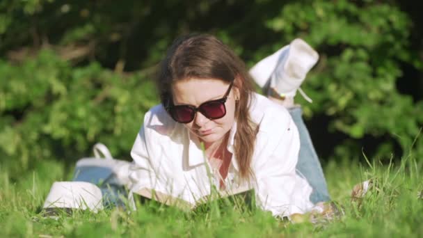 Erwachsene Frau mittleren Alters mit Sonnenbrille liegt mit Buch auf grünem Gras — Stockvideo