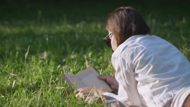 Женщина в солнечных очках лежит на зеленой траве в городском парке и читает книгу на открытом воздухе — стоковое видео