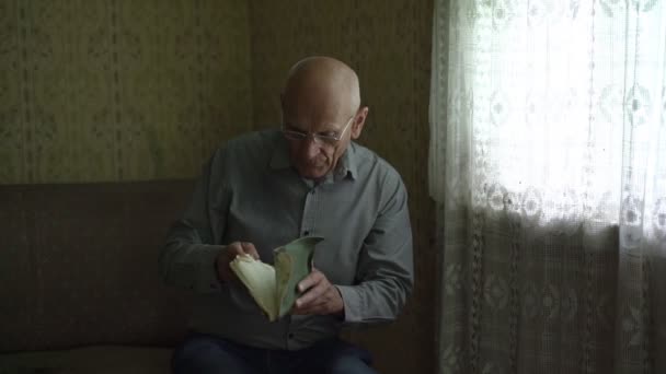 Homme âgé chauve en lunettes prendre livre de canapé et coup aux pages poussiéreuses. — Video