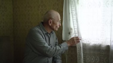 Gömlekli yaşlı adam perdeyi kaldırır ve pencereden dışarı bakar.