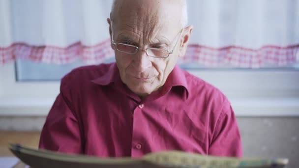 Staruszek w czerwonej koszuli i okularach czyta gazety w domu. — Wideo stockowe