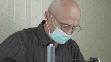 Gözlüklü ve mavi maskeli yaşlı adam eski makineye dikiş dikiyor.